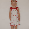 Young girl wearing a white dress with red accents and cherry pattern against a plain wall.