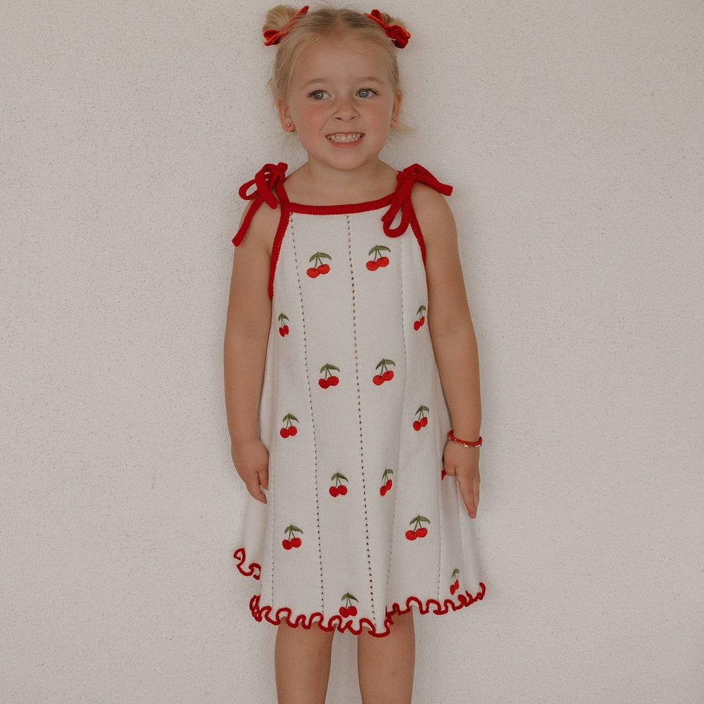 Young girl wearing a white dress with red accents and cherry pattern against a plain wall.