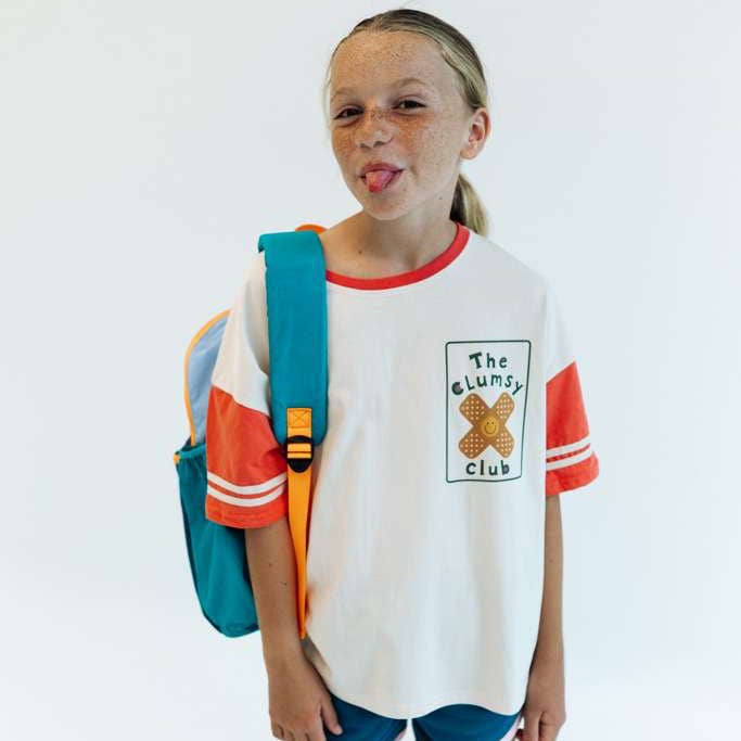A young girl with freckles wearing a white ringer t-shirt with orange and teal color-blocked sleeves and a graphic on the chest that reads 'The Clumsy Club'. She is carrying a teal and orange backpack against a plain white background.
