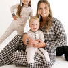 A woman and two children wearing matching patterned pajamas pose together on a clean white studio background. The woman wears a black-and-white gingham long-sleeve top and gingham pants, while the children wear light cream pajamas with small dark motifs.