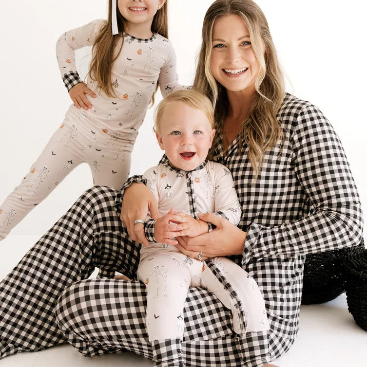 A woman and two children wearing matching patterned pajamas pose together on a clean white studio background. The woman wears a black-and-white gingham long-sleeve top and gingham pants, while the children wear light cream pajamas with small dark motifs.